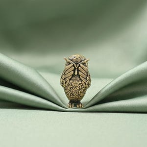 May include: A small, antique-style brass owl figurine with intricate engraved feather and floral patterns. The owl is depicted with its wings raised to cover its eyes, showcasing a detailed metallic texture and a warm, burnished gold finish.