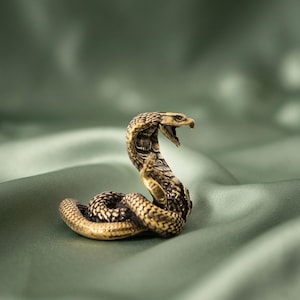 Puede incluir: Una pequeña figura de latón detallada de una cobra en posición enroscada y erguida, con la capucha desplegada y la boca abierta mostrando colmillos afilados. La superficie metálica presenta un patrón de escamas texturizado con reflejos.