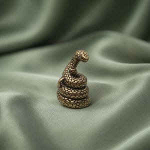 May include: A small, coiled brass snake figurine with a textured, scale-like surface. The snake is depicted in an upright, alert posture with its head raised and curved forward, showcasing a detailed metallic finish and a compact, rounded form.