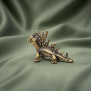 May include: A small, metallic bronze-toned figurine of a creature resembling a lizard or dragon. The figure features a series of sharp, pointed spikes running along its back and head, with a smooth, curved tail and four short legs.