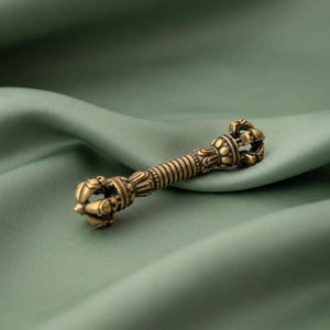 May include: A metallic gold-toned vajra ritual object featuring a central ribbed handle flanked by symmetrical, multi-pronged ends with intricate, detailed carvings and a weathered, antique-style finish throughout the entire metal surface.