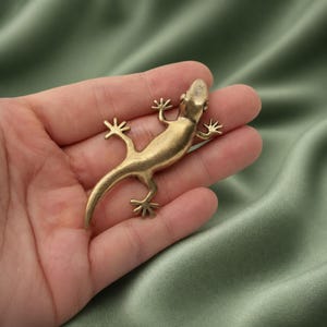 May include: A brass-toned metal lizard figurine with a smooth, matte finish. The lizard is depicted in a crawling position with four splayed feet, a long slender tail, and a rounded head. The surface of the metal shows subtle variations in color.