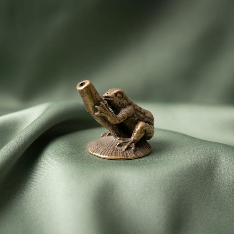K&ouml;nnte beinhalten: Eine kleine, metallische, bronzefarbene Froschfigur, die auf einer runden, strukturierten Basis sitzt. Der Frosch ist so dargestellt, dass seine Vorderbeine ein hohles, zylindrisches Rohr umschlie&szlig;en, das aus der Mitte der Basis ragt.