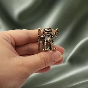 Puede incluir: Una pequeña figura de latón de estilo antiguo que representa una rana con una capa con capucha, sosteniendo un bastón alto y texturizado en una mano y un libro abierto en la otra. El metal tiene una pátina oscura y desgastada.