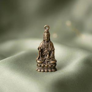 Può includere: Un piccolo ciondolo in ottone in stile antico raffigurante una figura seduta con vesti, poggiata su una base a forma di loto. La figura ha una mano sollevata e tiene un piccolo vaso. Un occhiello circolare è fissato sulla parte superiore.