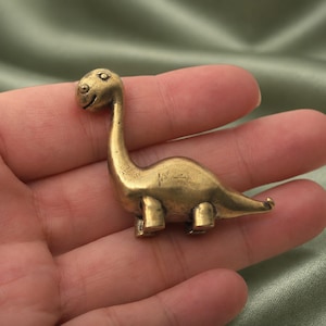 May include: A small, solid brass figurine shaped like a long-necked dinosaur. The metal has a polished, golden-brown finish with a smooth, rounded body, a curved neck, a small head with etched eye details, and four short, sturdy legs.