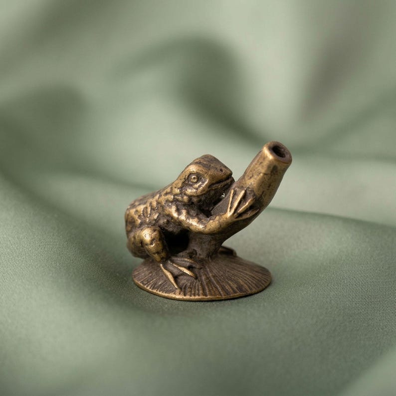 K&ouml;nnte beinhalten: Eine kleine, antik wirkende Messingfigur eines Frosches auf einem runden Sockel. Der Frosch hat eine texturierte Haut und h&auml;lt ein hohles, zylindrisches Rohr, das schr&auml;g nach oben ragt und als kleiner Halter oder St&auml;nder dient.