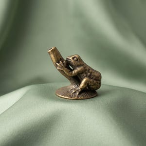 K&ouml;nnte beinhalten: Eine kleine Messingfigur im Antik-Stil, die einen Frosch auf einem strukturierten, zylindrischen Ast darstellt. Der Frosch hat eine detaillierte Hautstruktur und sitzt auf einem runden, geriffelten Sockel. Das Metall hat eine dunkle Patina.