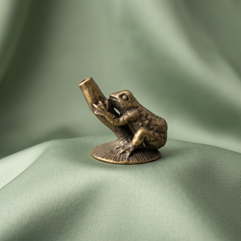 K&ouml;nnte beinhalten: Eine kleine Messingfigur im Antik-Stil, die einen Frosch auf einem strukturierten, zylindrischen Ast darstellt. Der Frosch hat eine detaillierte Hautstruktur und sitzt auf einem runden, geriffelten Sockel. Das Metall hat eine dunkle Patina.