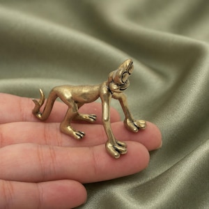 Könnte beinhalten: Eine kleine, metallische, messingfarbene Figur eines stilisierten Wesens mit einem langen, geschwungenen Schwanz, verlängerten Gliedmaßen und markanten, runden Augen. Die Figur ist in einer vierbeinigen Haltung mit glatter Oberfläche dargestellt.