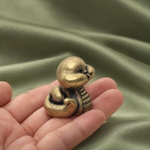 以下が含まれることがあります： 丸みを帯びた頭部と胴体に刻まれた細かな線が特徴的な、真鍮製の小さな蛇の置物。アンティーク調の仕上げが施されており、金属特有の光沢のある金茶色の表面には、溝の部分に暗い色合いのアクセントが加わっています。