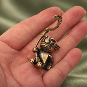 May include: A small, antique-style brass figurine of a seated creature with large, wide-set eyes and a downturned mouth. The figure holds a long, curved staff that arches over its head, featuring a serrated, gear-like top edge and a dark patina.