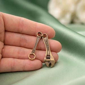 May include: A small metallic pendant shaped like two miniature tools, a fixed wrench and an adjustable wrench, linked together. The adjustable wrench features the text "SILVER 925" engraved on its handle, while the fixed wrench has "925" on it.