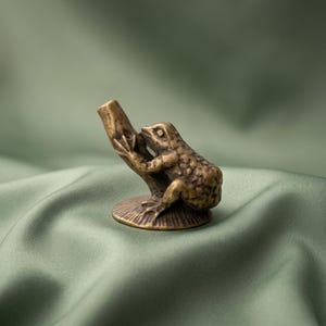 K&ouml;nnte beinhalten: Eine kleine, antik wirkende Messingfigur eines Frosches, der auf einem strukturierten, zylindrischen Sockel sitzt. Der Frosch ist mit detaillierter Hautstruktur, Schwimmh&auml;uten und einer fokussierten Haltung auf einer runden Metallplatte dargestellt.