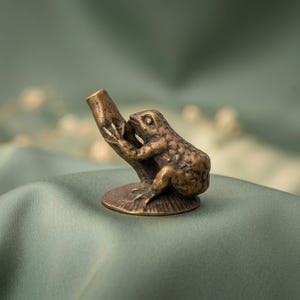 K&ouml;nnte beinhalten: Eine kleine, antik wirkende Metallfigur in Bronzet&ouml;nen, die einen Frosch auf einem strukturierten Ast darstellt. Der Frosch ist mit detaillierter Hautstruktur geformt und sitzt auf einem runden, geriffelten Sockel. Das Objekt hat eine hohle, abgewinkelte &Ouml;ffnung an der Spitze des Astes, was auf ein funktionales Design zum Halten eines d&uuml;nnen Gegenstands hindeutet.
