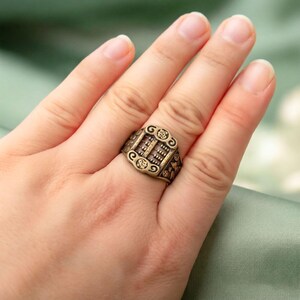 May include: A bronze-toned metal ring featuring a miniature abacus design on the face. The band displays intricate embossed details, including small circular motifs and scrollwork patterns surrounding the central abacus frame.