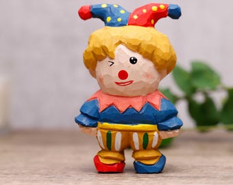 Peut inclure: Figurine de clown en bois sculpté à la main. Le clown a un nez rouge, un œil qui cligne et une bouche souriante. Il porte un chapeau de bouffon bleu et rouge, un col rose, un haut bleu et un pantalon jaune à rayures vertes et blanches. Les chaussures du clown sont rouges et bleues.