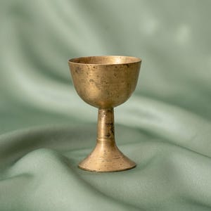 May include: A small, weathered brass chalice featuring a smooth, rounded bowl, a slender central stem, and a flared circular base. The metal surface displays a muted, aged patina with subtle dark spots and variations in the golden-brown tone.