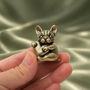 May include: A small, antique-style brass figurine of a French Bulldog sitting upright. The dog has large, pointed ears, a wrinkled face, and is holding a smooth, rounded object against its chest. The metal has a weathered, golden-brown finish.