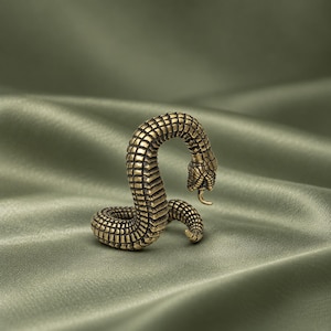 May include: A small, antique-style brass figurine of a coiled snake with a textured, scale-like surface. The snake is depicted in an upright, alert position with its head curved downward and a forked tongue protruding from its mouth.