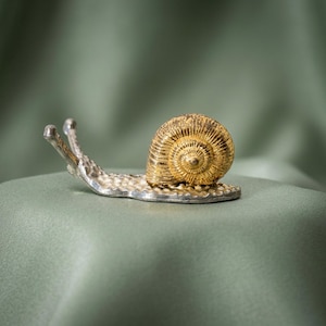 Pode incluir: Uma figura decorativa de caracol com um corpo de metal prateado de superfície texturizada e uma concha dourada em espiral proeminente. O caracol tem dois pedúnculos oculares estendidos e uma forma detalhada, contrastando o prata e o dourado.