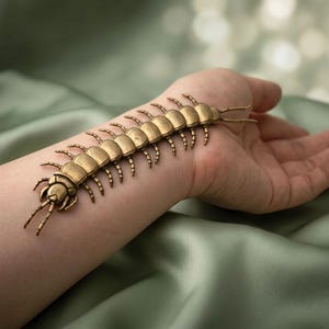 May include: A metallic gold-toned centipede ornament featuring a segmented body with multiple curved legs and two long antennae extending from the head. The insect-shaped piece has a textured, antique-style finish with visible joints.