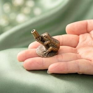 K&ouml;nnte beinhalten: Eine kleine, antik wirkende Metallfigur in Bronzefarbe, die einen Frosch auf einer runden, strukturierten Seerosenblatt-Basis darstellt. Der Frosch sitzt und h&auml;lt ein hohles, zylindrisches, r&ouml;hrenf&ouml;rmiges Objekt, das nach oben ragt.