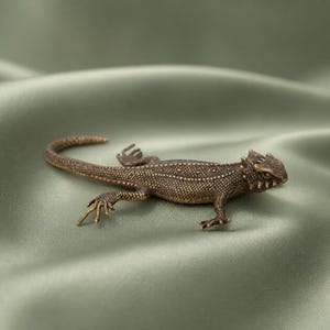 May include: A small, detailed bronze-toned metal lizard figurine featuring a textured, scaly surface, a long curved tail, and distinct spikes around its head. The lizard is depicted in a crawling position with four legs extended outward.
