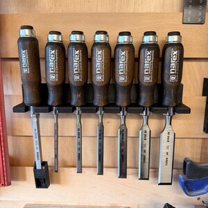 May include: Seven wood chisels with dark wooden handles and silver blades, displayed on a black holder. Each chisel is labelled "narex Made in Czech Republic" with size markings. The chisels are mounted on a wooden surface.