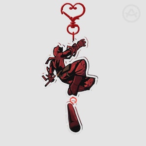 Könnte beinhalten: Ein roter Schlüsselanhänger mit einer Cartoon-Illustration eines Superhelden in dynamischer Pose. Der Schlüsselanhänger hat einen herzförmigen Verschluss und einen zylindrischen Anhänger. Der Superheld trägt einen roten Anzug mit schwarzen Akzenten und hält eine Waffe.