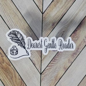 May include: A white sticker with a black illustration of a quill pen, inkwell, and the words "Dearest Gentle Reader" in elegant script. The sticker is placed on a wooden surface with a chevron pattern.