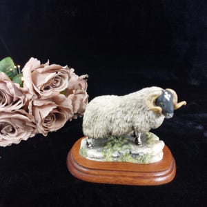 May include: A white sheep figurine with black markings on its face and horns. The sheep is standing on a green and brown base with a wooden base.
