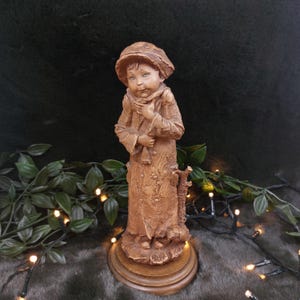 Può includere: Una statuetta di legno di un giovane ragazzo che indossa un cappello e un lungo cappotto. La statuetta è in piedi su una base di legno.