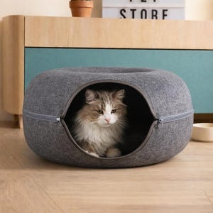 Puede incluir: Una cama gris para gatos con una abertura con cremallera, que alberga un gato esponjoso con pelaje marrón y blanco. La cama es de forma ovalada y está sobre un suelo de madera. Un letrero que dice "HAPPY PET STORE" está en el fondo.