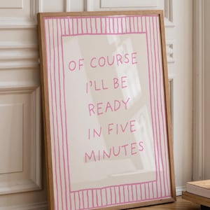May include: A framed art print with the text "OF COURSE I'LL BE READY IN FIVE MINUTES" in pink lettering. The print has a pink striped border and a light wood frame. The artwork is leaning against a white door.