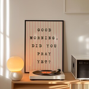 May include: A wooden cabinet with a record player, a lamp, and a framed poster that reads "GOOD MORNING, DID YOU PRAY YET?". The cabinet has a collection of vinyl records. A black and white speaker is on the right.
