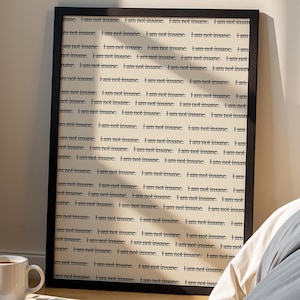 May include: A framed print with the repeated text "I am not insane" in black font. A white mug sits on a stack of books, and a black lamp is in the background. The scene is set in a bedroom with a bed and a window.