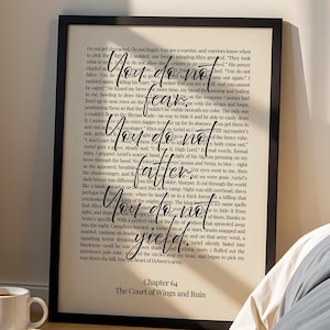 Puede incluir: Una impresión enmarcada con el texto "You do not fear, falter, yield" en una escritura elegante. La impresión está apoyada contra una pared junto a una cama con ropa de cama gris. Una lámpara negra y una pila de libros con una taza de café están en primer plano.