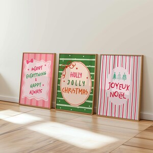 May include: Three framed Christmas art prints. The prints feature festive text in pink, green, and red, with phrases like "Merry Everything & Happy Always," "Holly Jolly Christmas," and "Joyeux No&euml;l." The frames are a natural wood color.