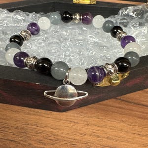 Può includere: Un braccialetto di perline con perline viola, bianche, grigie e nere. Presenta un ciondolo argentato a forma di pianeta. Il braccialetto è presentato all'interno di una scatola di legno.