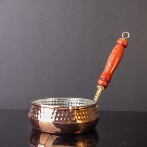 May include: A copper saucepan with a hammered finish and a wooden handle. The saucepan has a small spout on the side.