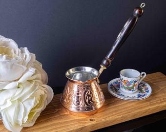 May include: A copper Turkish coffee pot with an ornate design and a wooden handle. The pot is sitting on a wooden surface next to a small white coffee cup with a floral design.