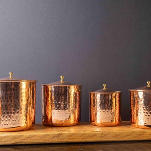 May include: Four copper canisters with a hammered finish and gold lids, arranged in a row on a wooden surface.