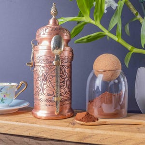 May include: A copper coffee pot with intricate floral designs and a brass handle. The pot has a lid with a finial and a spoon inside. A glass jar with a cork lid contains ground coffee beans. A wooden spoon is on the table next to the jar.