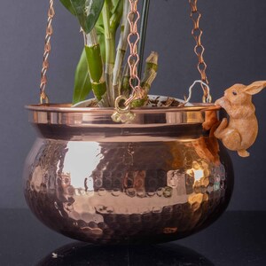 May include: A copper hanging planter with a hammered finish and a chain for hanging. A small brown rabbit figurine is attached to the side of the planter.