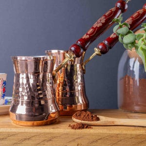 May include: Two hammered copper coffee pots with wooden handles, one filled with coffee grounds, on a wooden surface with a spoon and a small white cup.