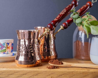 May include: Two hammered copper coffee pots with wooden handles, one filled with coffee grounds, on a wooden surface with a spoon and a small white cup.