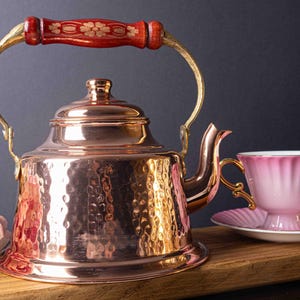 May include: A hammered copper tea kettle with a wooden handle and brass accents. The kettle is sitting on a wooden surface next to a pink teacup and saucer.