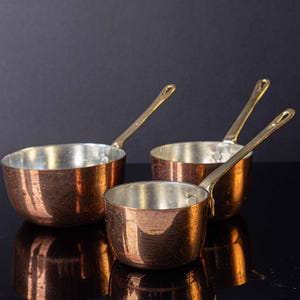 May include: Three copper saucepans with brass handles. The saucepans are nested together, with the smallest saucepan on top. The saucepans are all shiny and new.