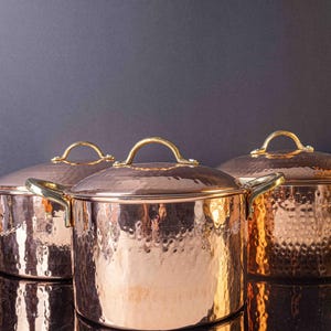 May include: Three copper pots with hammered surfaces and brass handles. The pots are arranged in a row, with the largest pot in the centre.